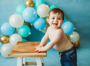 Baby in blue jeans poses on wooden table with turquoise and gold balloons in background during milestone photoshoot.
