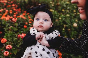 A baby wearing a polka dot outfit sits in a garden with orange flowers in the background.