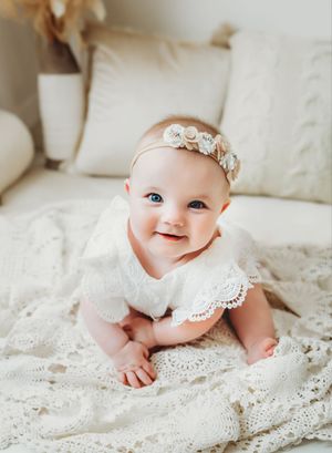 Baby wearing delicate floral headband lies on white lace blanket in sweet portrait series.