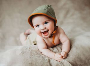 Baby wearing brown cap smiles while lying on soft beige fabric in candid photo series.