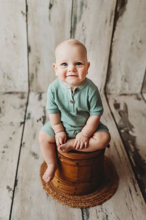 A baby in a mint green outfit sits on a wooden stool against a rustic white wooden plank backdrop.