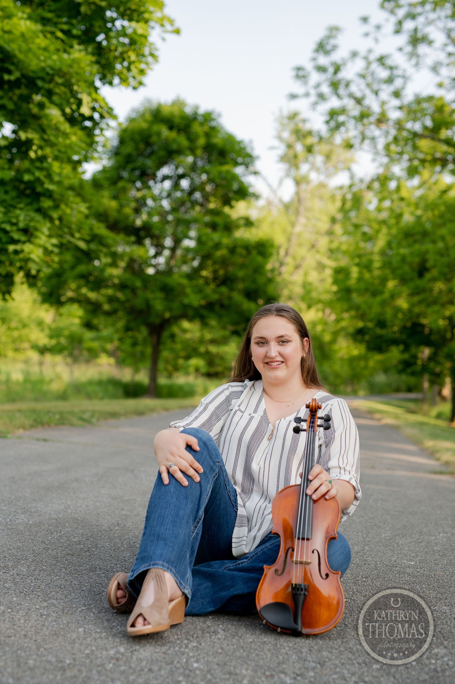Violinist sits on path holding instrument against backdrop of summer trees.