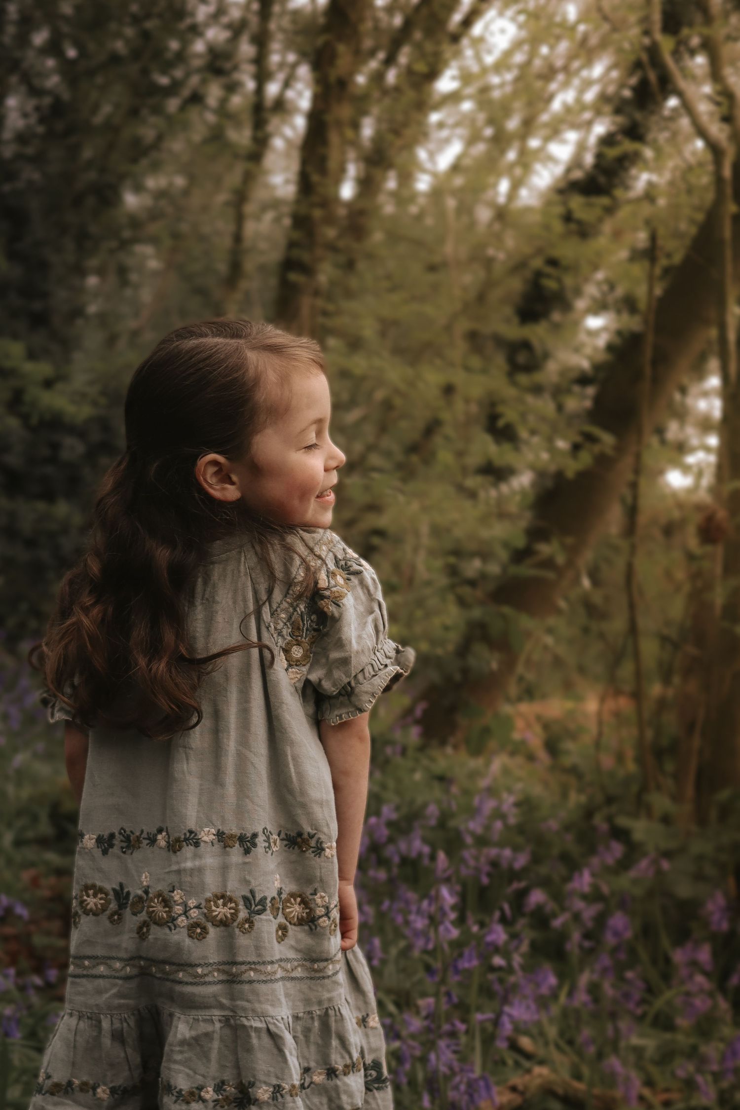 A child in a vintage-style embroidered dress stands in a forest clearing amongst the bluebells