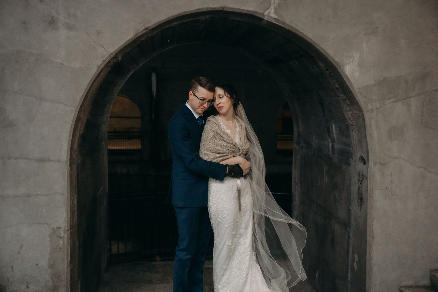 A romantic wedding portrait of a couple embracing under a stone archway.