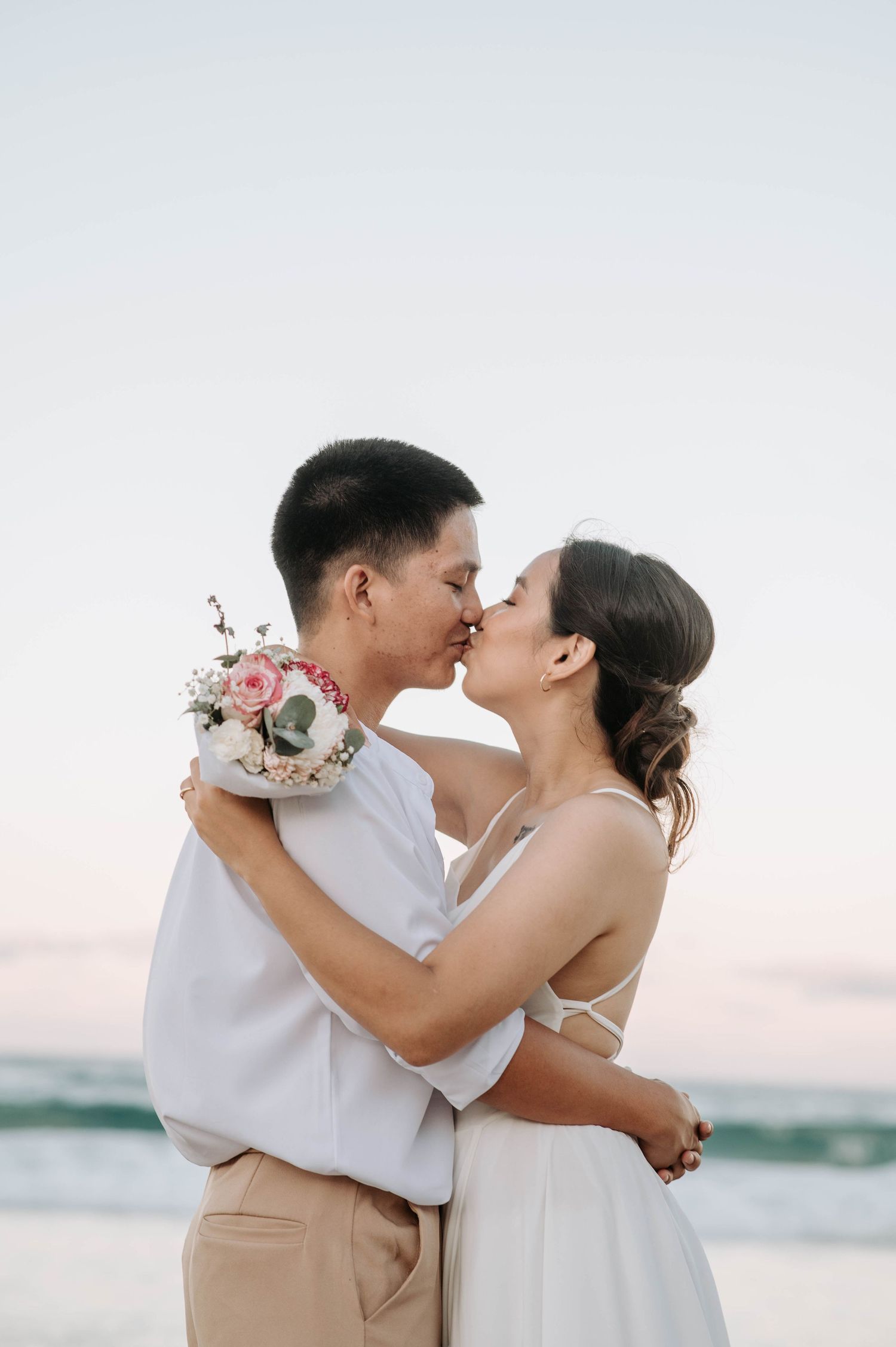 A romantic kiss between a couple in wedding attire on the beach at sunset.
