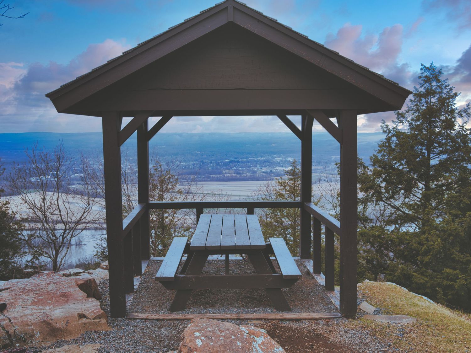 Wooden gazebo with picnic table overlooking a scenic valley and river at dusk in winter.
