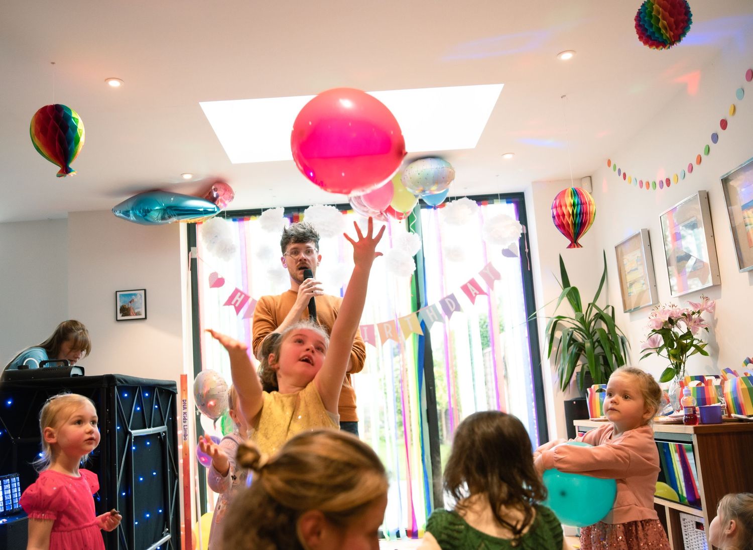 Children reach up excitedly to catch colorful balloons at an indoor birthday party decorated with streamers and banners.