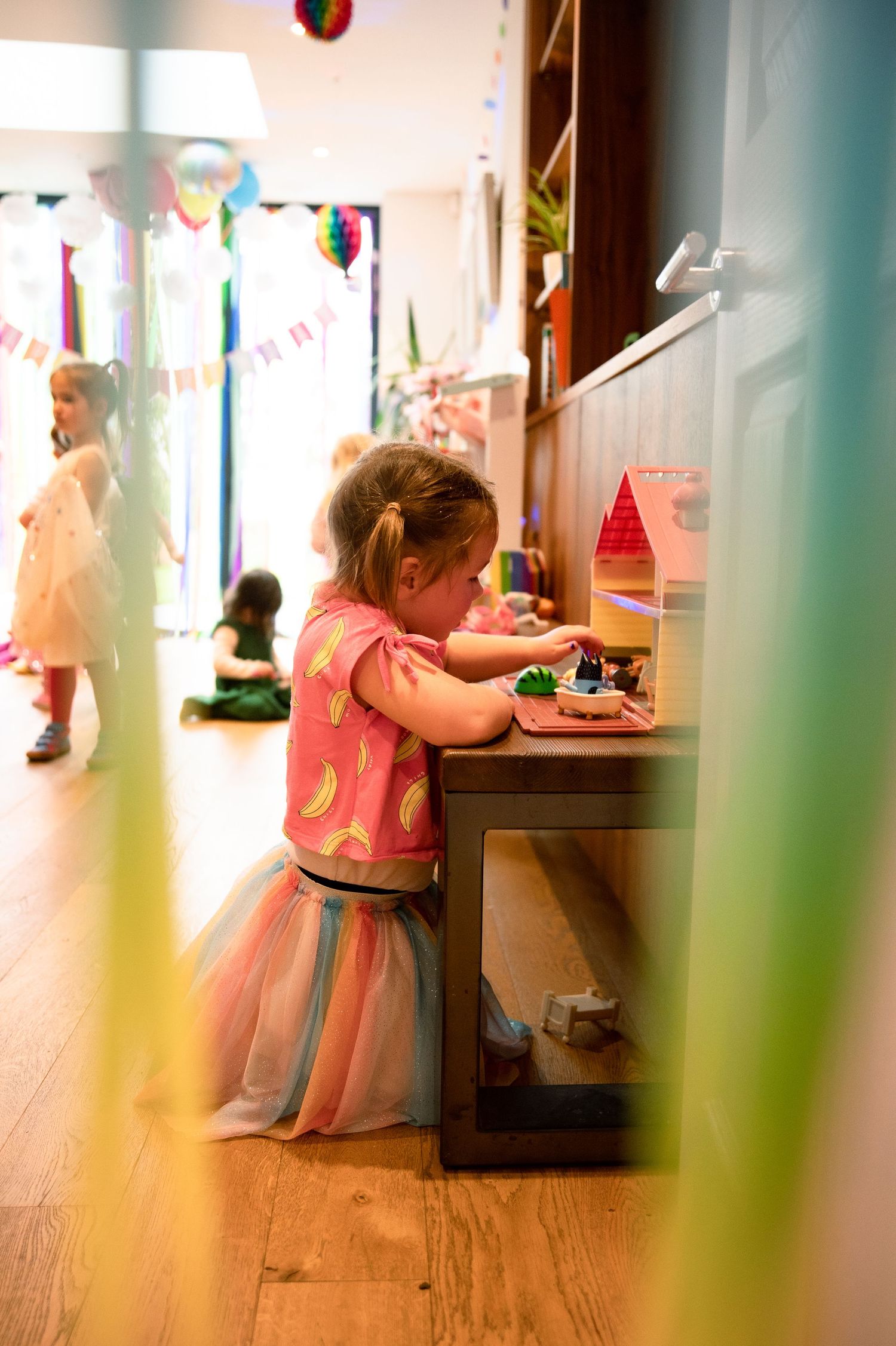 Small child in a pink outfit plays at a wooden cabinet while party decorations and balloons hang in the background.
