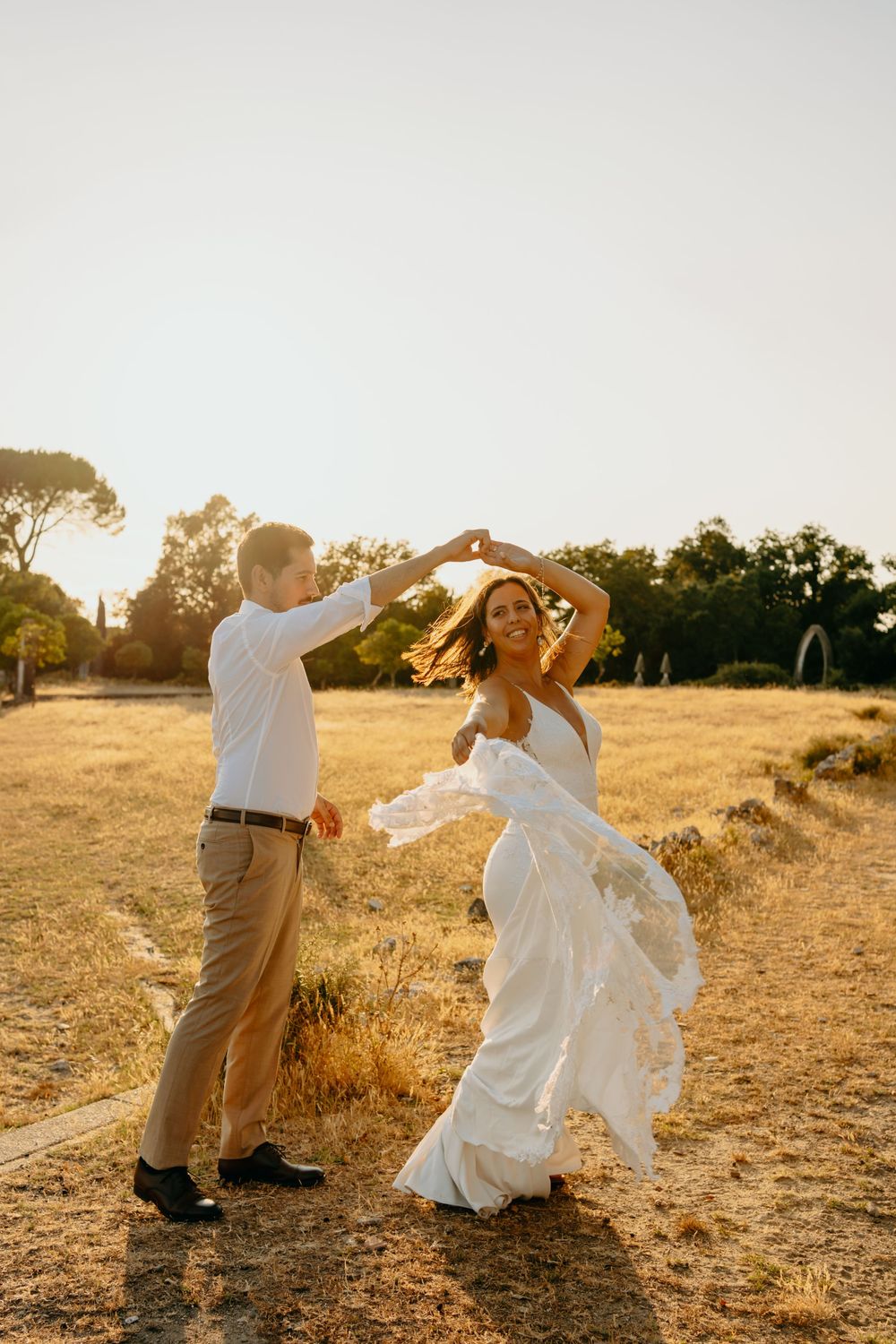 Couple dances together in a sun-drenched field at golden hour, bride wearing flowing white wedding dress.