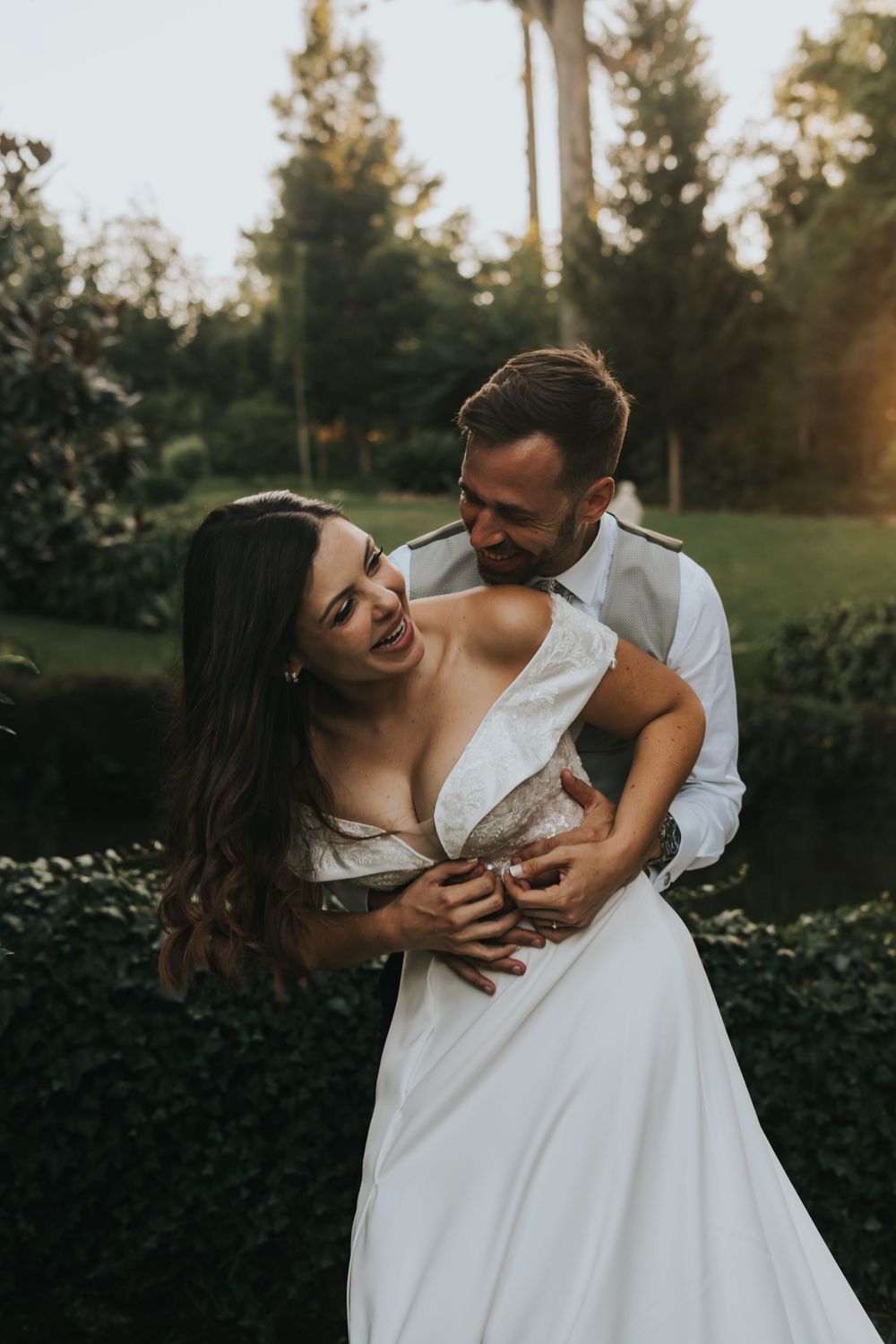 A romantic couple embraces in a garden at sunset, the bride wearing a flowing white dress as they share an intimate moment.