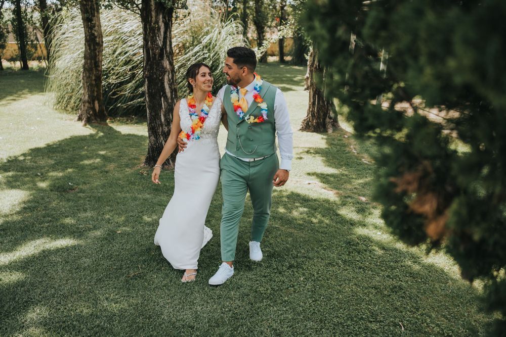 A couple in wedding attire embraces outdoors on a sunny day beneath tall trees in a parklike setting.