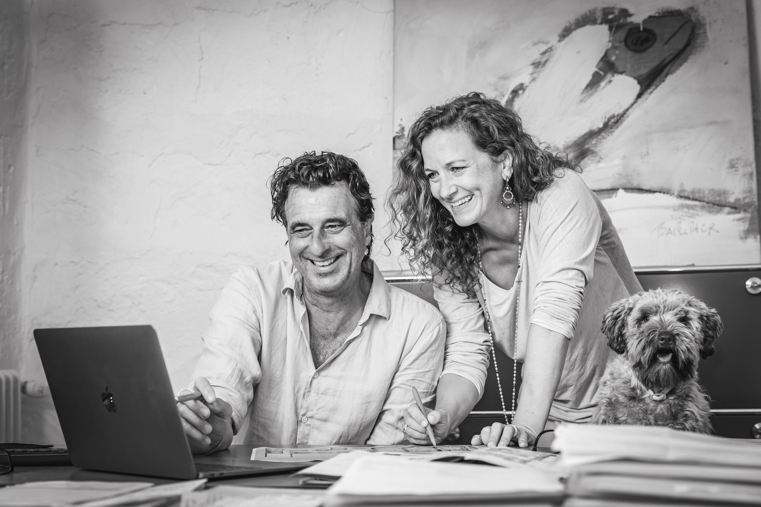 Black and white candid portrait of a smiling couple working at a desk with a laptop and their small dog in a Mallorca home.