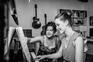 Black and white photo of a young woman smiling and sitting at a piano with her female piano teacher with guitars in the background.