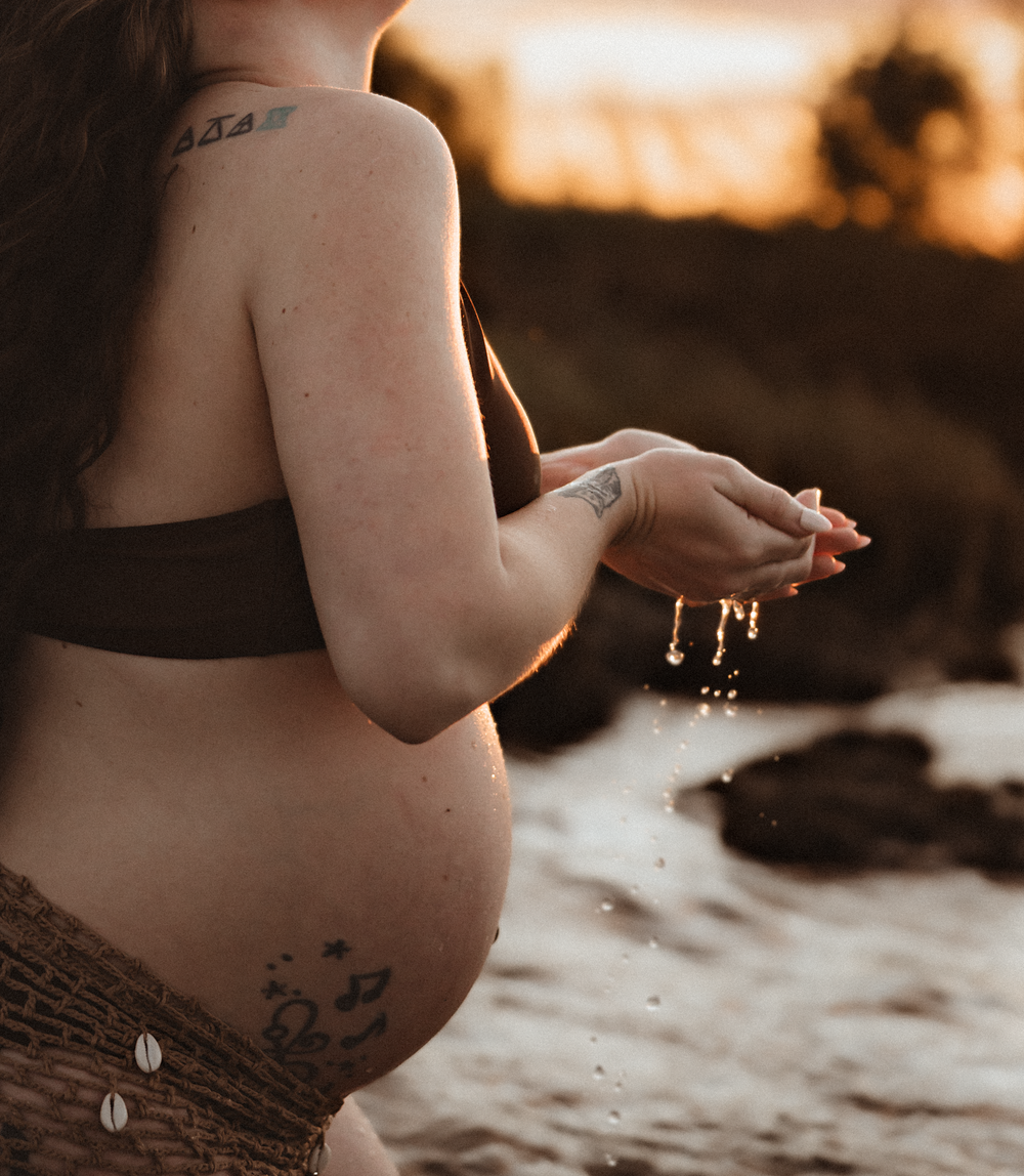 Pregnant woman in brown bikini top with tattoos letting sand fall from hands at sunset beach.