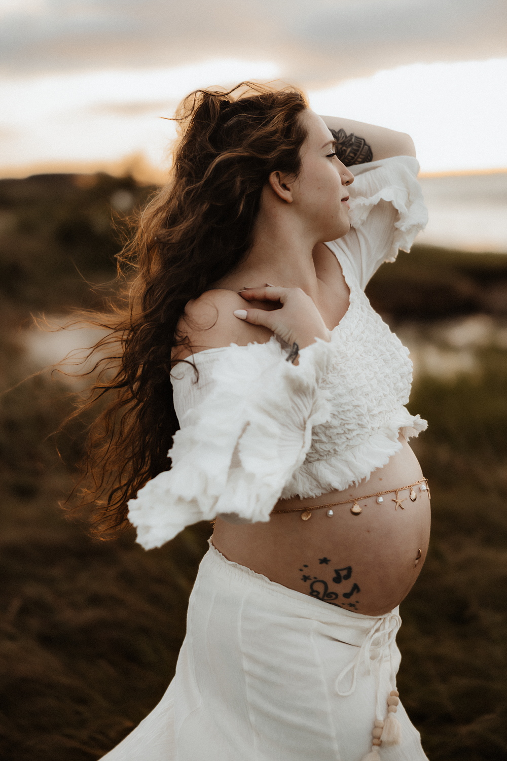 Pregnant woman in white crop top and skirt with belly art poses outdoors at sunset with flowing hair.