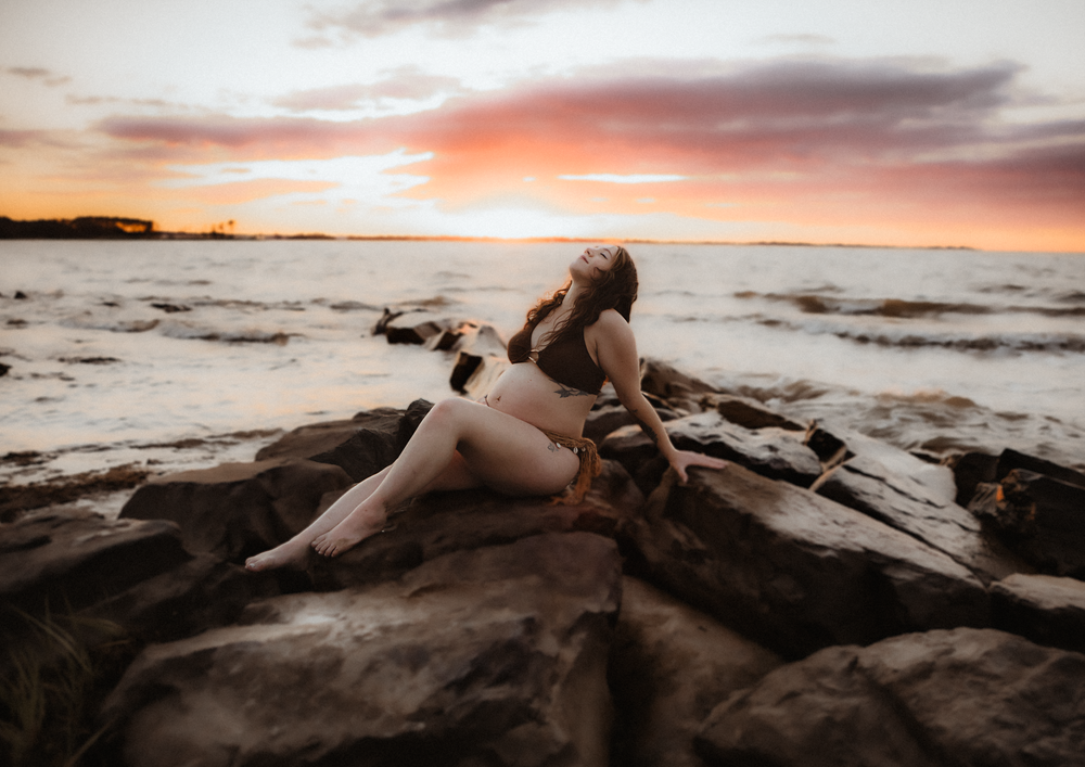 Woman poses on rocky shoreline at sunset with cat, warm orange sky reflecting over calm ocean waters.