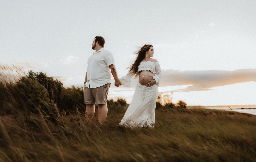 Pregnant couple holding hands in a golden field at sunset, woman in white dress cradling baby bump.