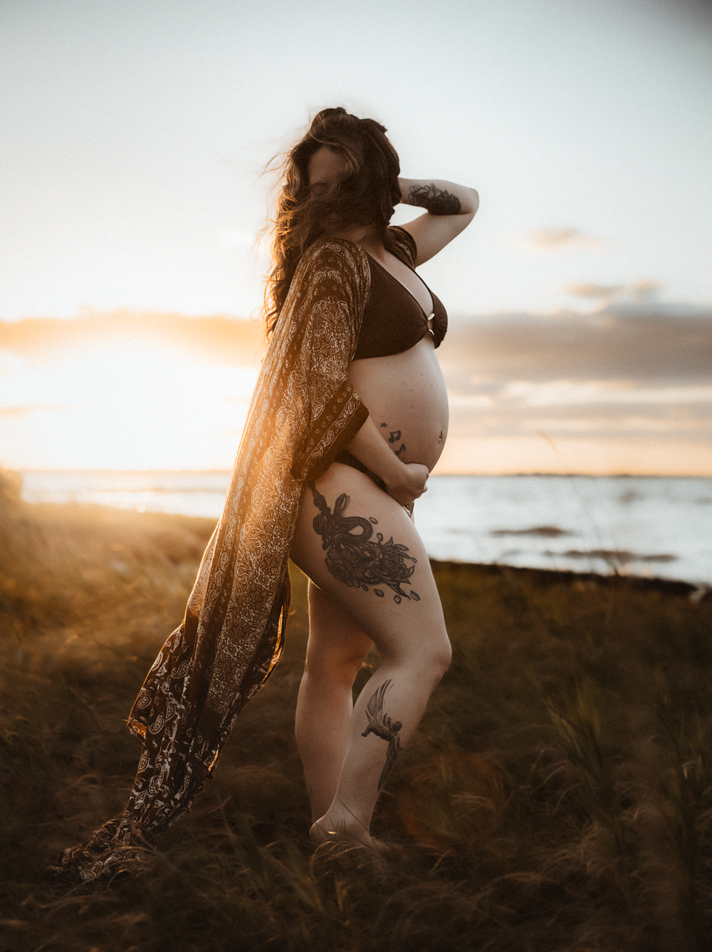 Pregnant woman with tattoos poses at golden hour sunset on beach wearing black bikini top and floral kimono.