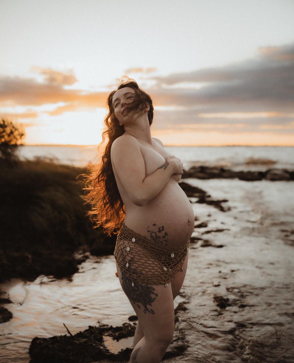 Pregnant woman in mermaid costume poses on rocky beach at golden sunset with long flowing hair.