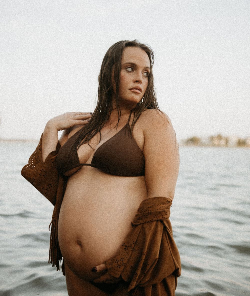 A series of intimate maternity photos showing a pregnant woman in a brown sweater posing in the ocean at sunset.