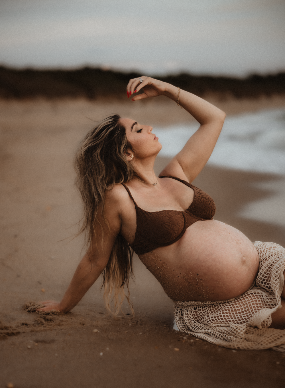 Pregnant woman in brown bikini top poses gracefully on sandy beach, with flowing hair and mesh skirt, during golden hour.