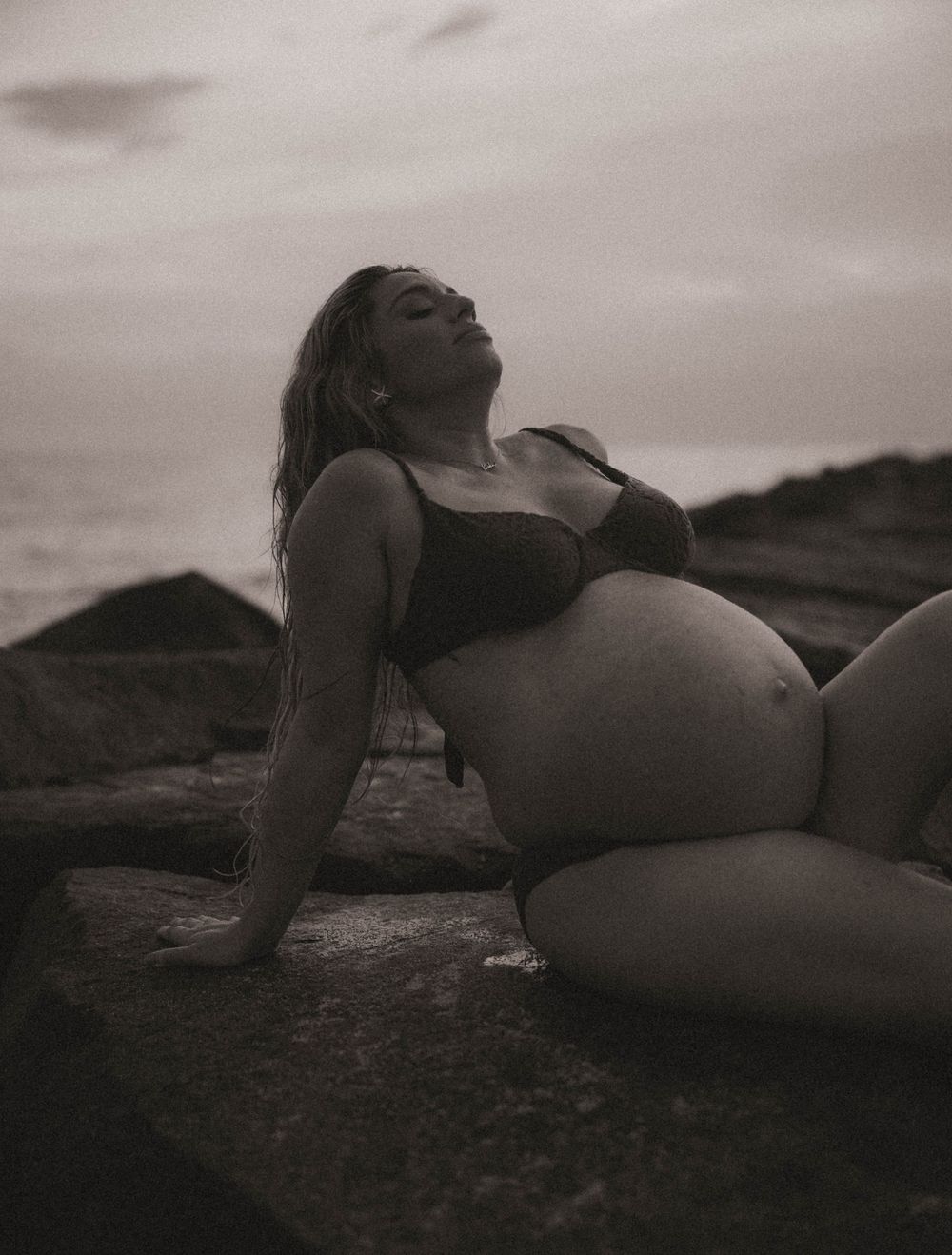 Black and white maternity photo of pregnant woman in bikini posing on rocks by the ocean at dusk.
