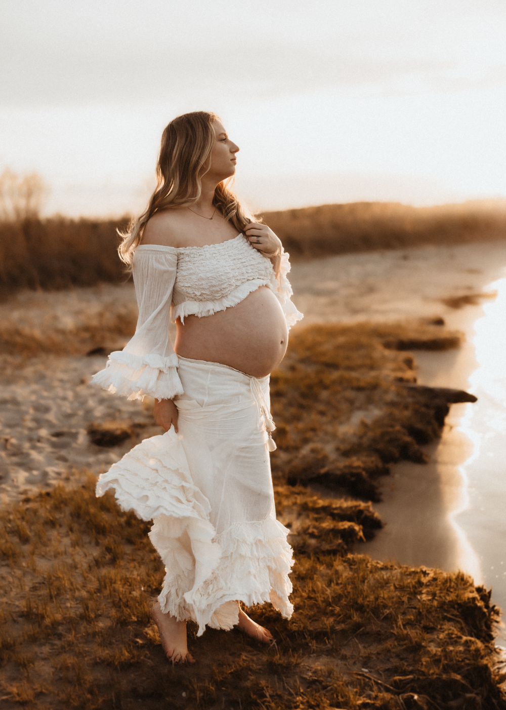 Pregnant woman in white boho dress posing outdoors at golden hour near a serene riverbank.