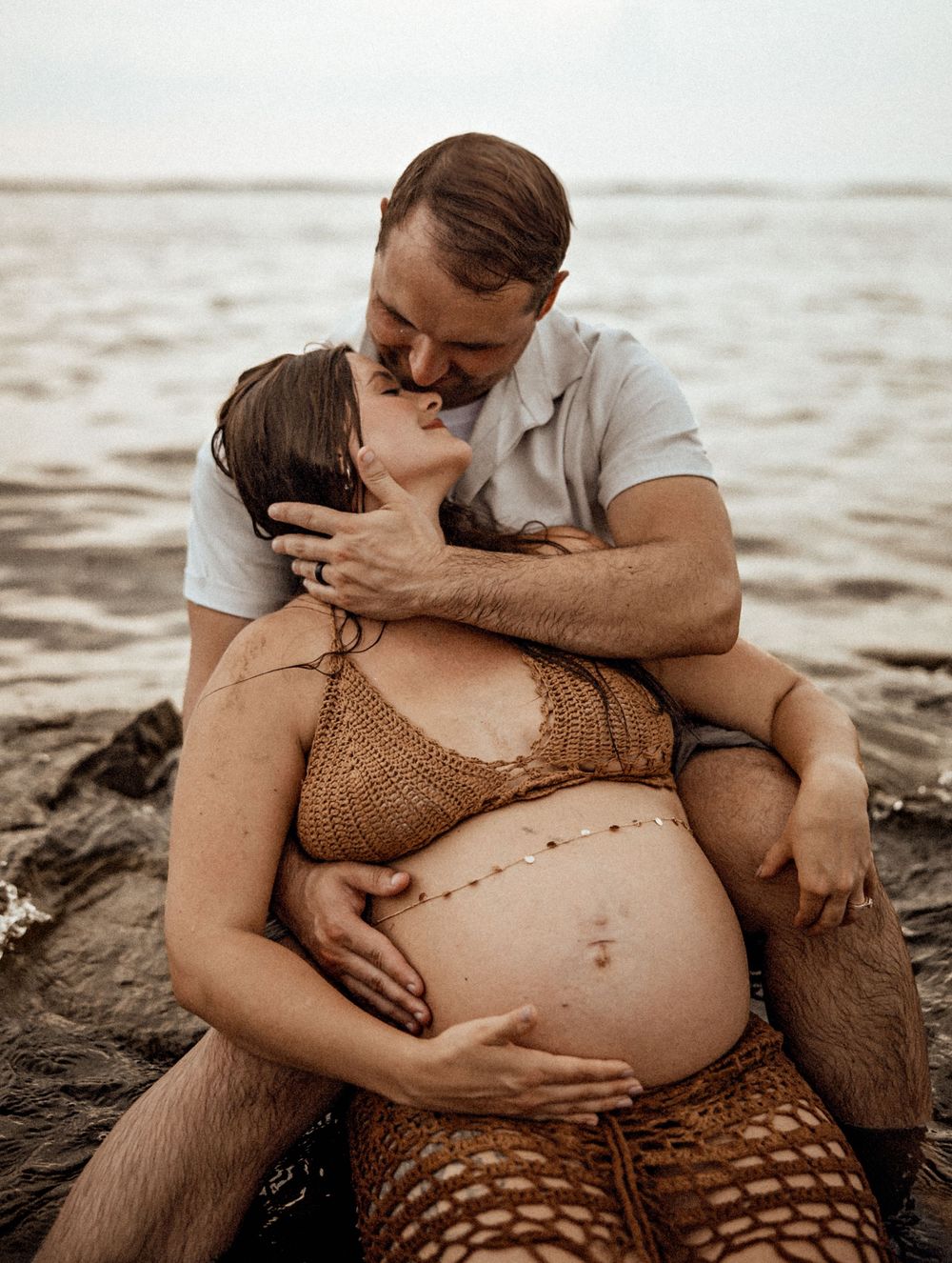Intimate maternity beach photo showing an expectant couple sharing a tender moment on rocky shore.