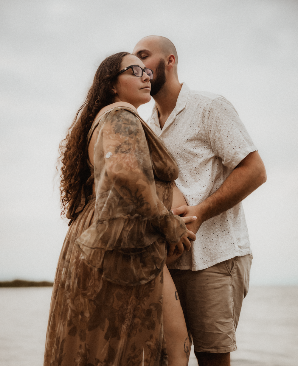 A couple shares a tender moment on the beach, with the man kissing his pregnant partner's head near the ocean.