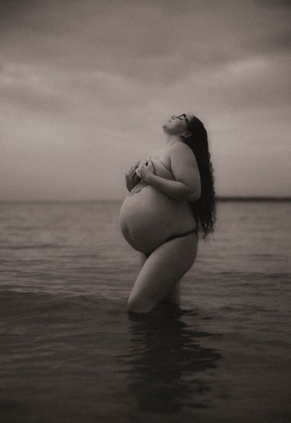Black and white photo of a pregnant woman standing in ocean water, head tilted back, long hair flowing.