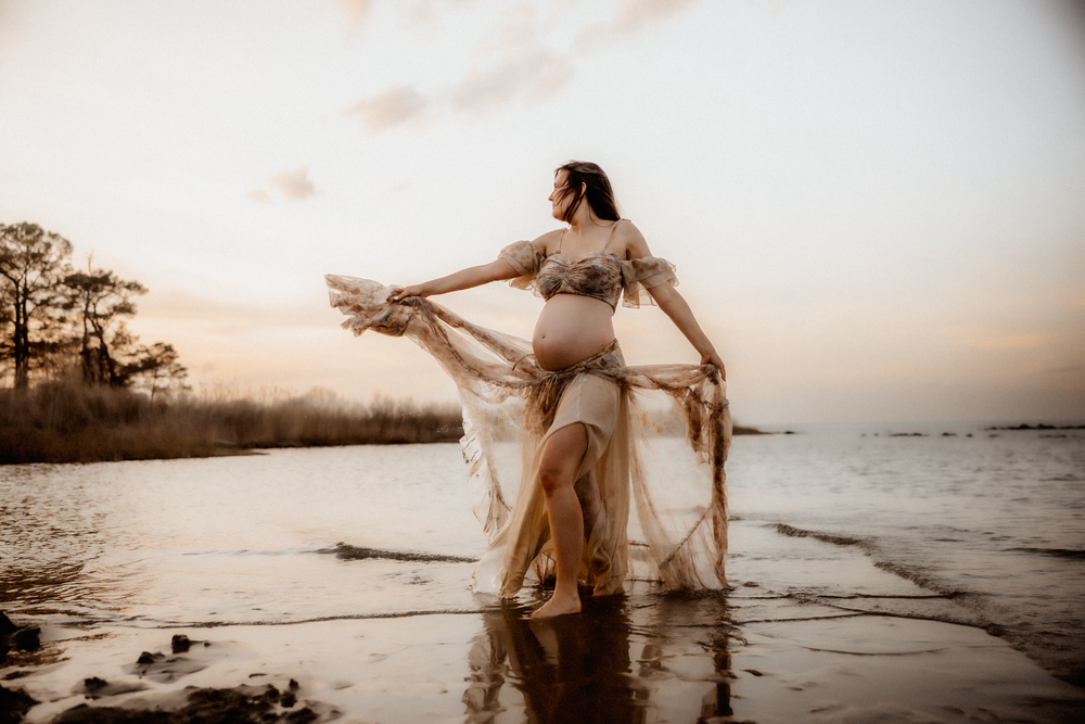Pregnant woman in flowing boho gown poses gracefully by the water at golden hour sunset.