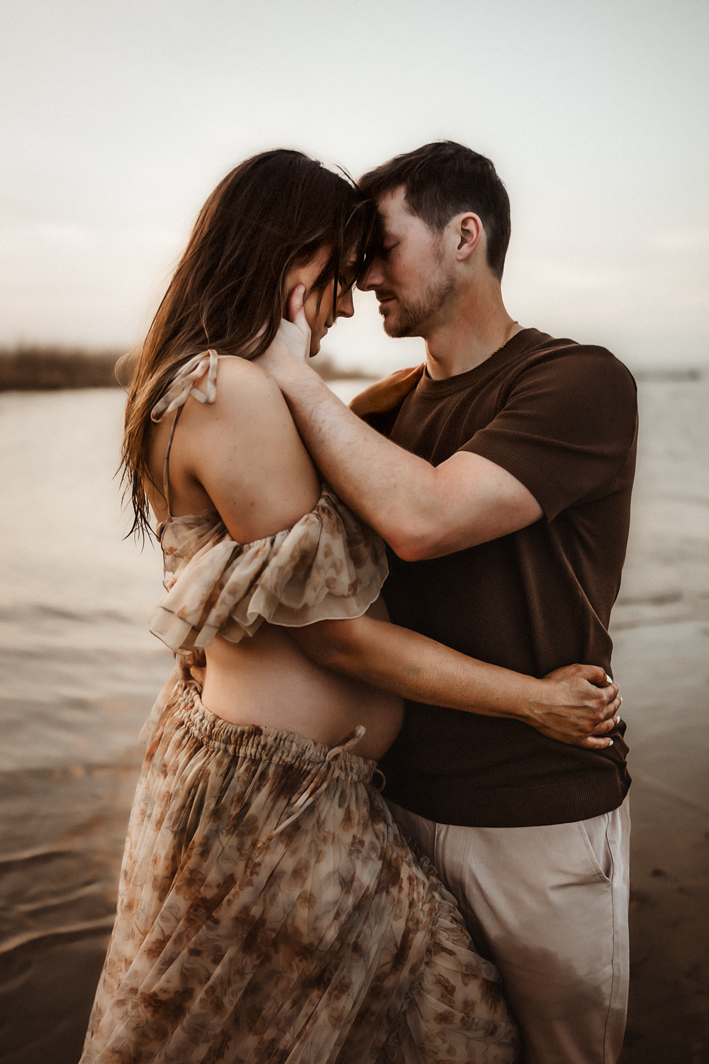 A couple shares an intimate moment on a beach at sunset, with the woman showing a baby bump in a floral dress.