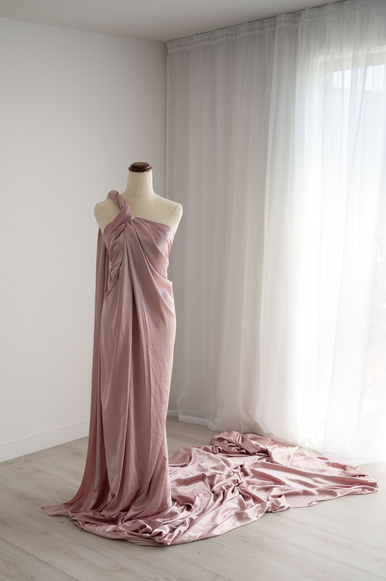 Dusty rose colored flowing fabric dress on mannequin with long train against white curtained window.