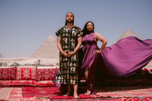 A couple poses against the backdrop of Egyptian pyramids on a red carpet with flowing purple fabric.