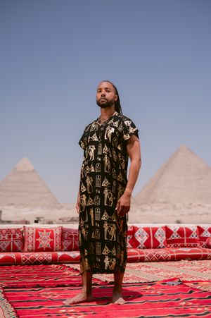 Person in patterned dress standing on red carpet with pyramids of Giza in background.