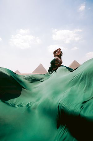 A flowing green dress billows dramatically against the backdrop of Egyptian pyramids under a blue sky.