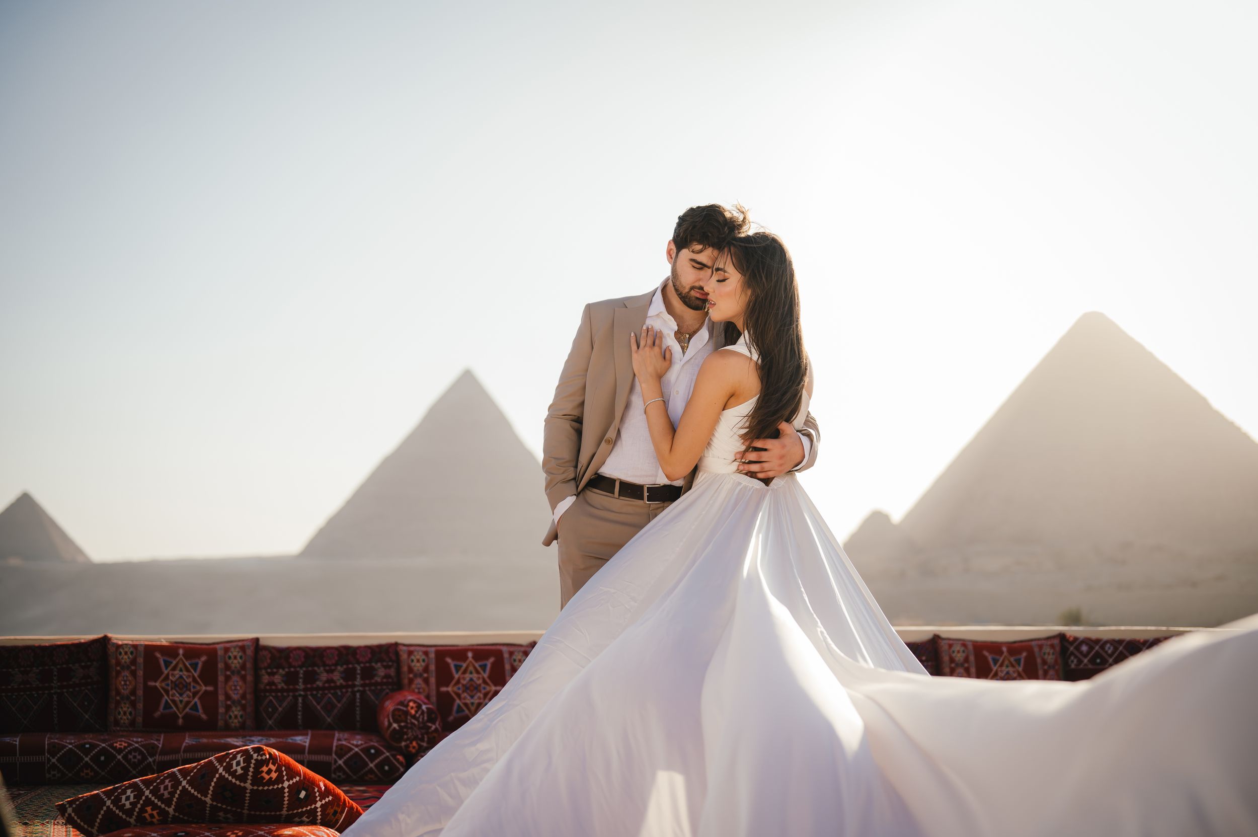 Romantic couple in wedding attire embracing with pyramids of Giza in background.