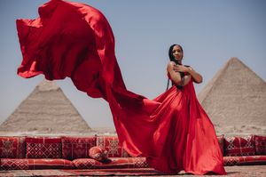 A red flowing dress billows dramatically against the backdrop of the ancient Egyptian pyramids at Giza.