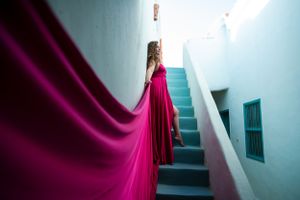 Figure in flowing magenta dress poses on turquoise staircase in architectural setting.