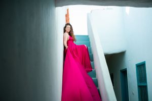 Person in vibrant pink gown standing on blue-walled staircase with dramatic fabric flow.