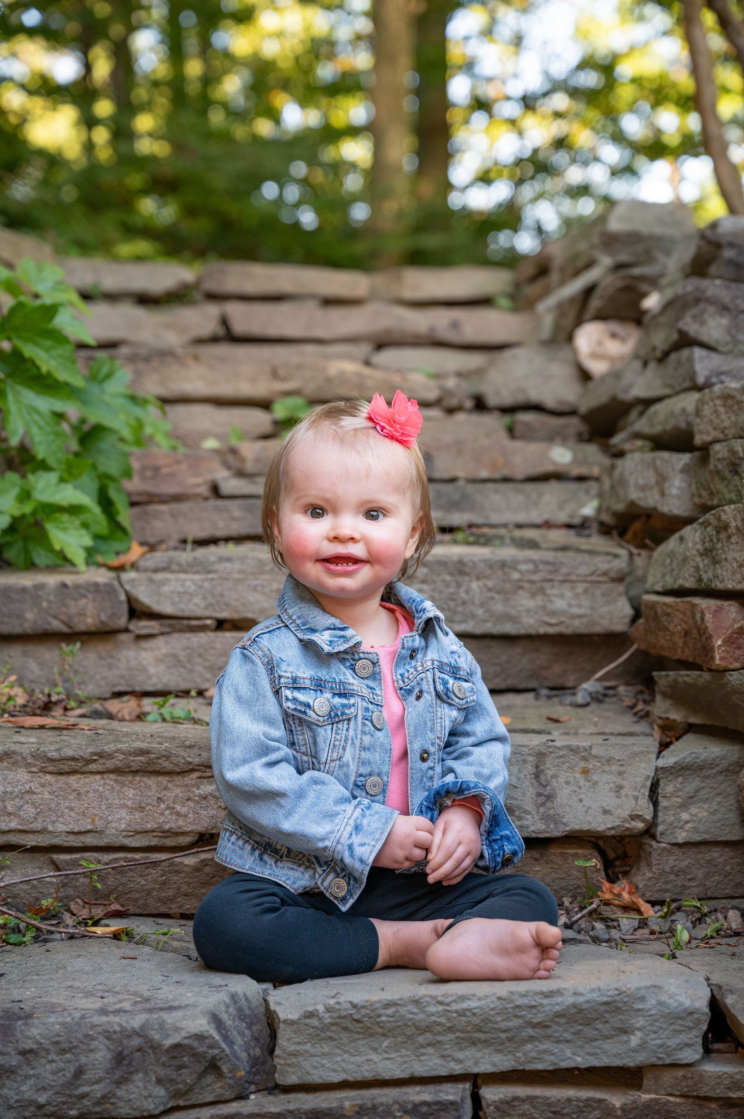 Small child wearing denim jacket sits on stone steps surrounded by greenery in outdoor portrait setting.