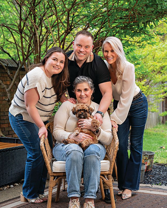 A family group poses together on a patio with their small brown dog in a wooden chair surrounded by greenery.