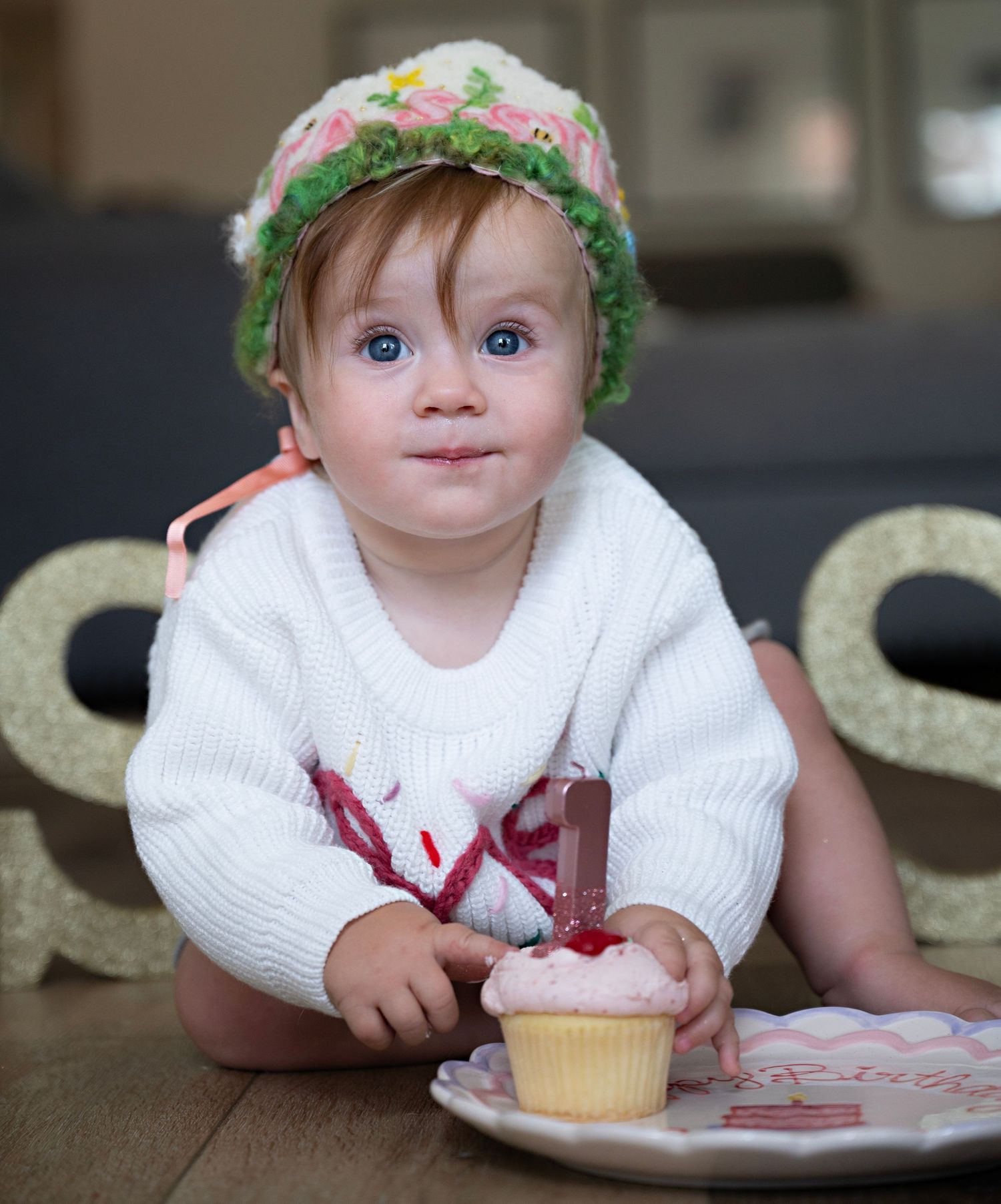 Baby wearing a knit white sweater and floral hat sits by a cupcake on a wooden table.