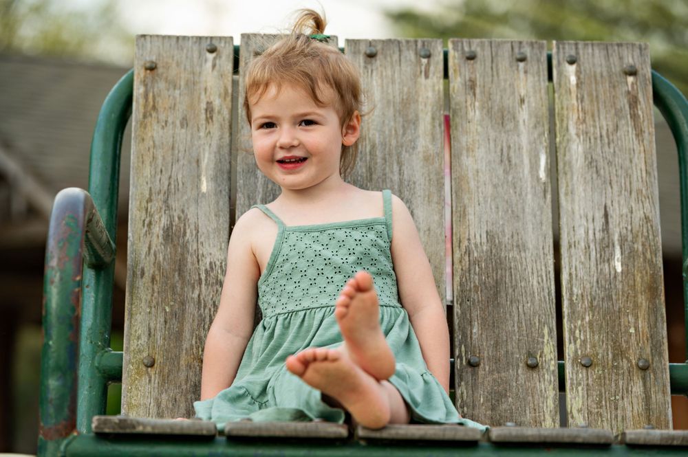 Smiling toddler in mint green dress sitting on rustic wooden bench  during a family portrait session on a summer evening.