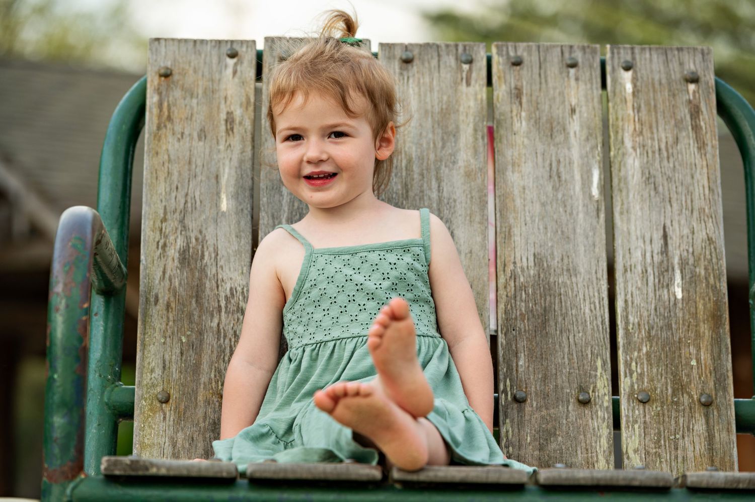 Smiling toddler in mint green dress sitting on rustic wooden bench  during a family portrait session on a summer evening.