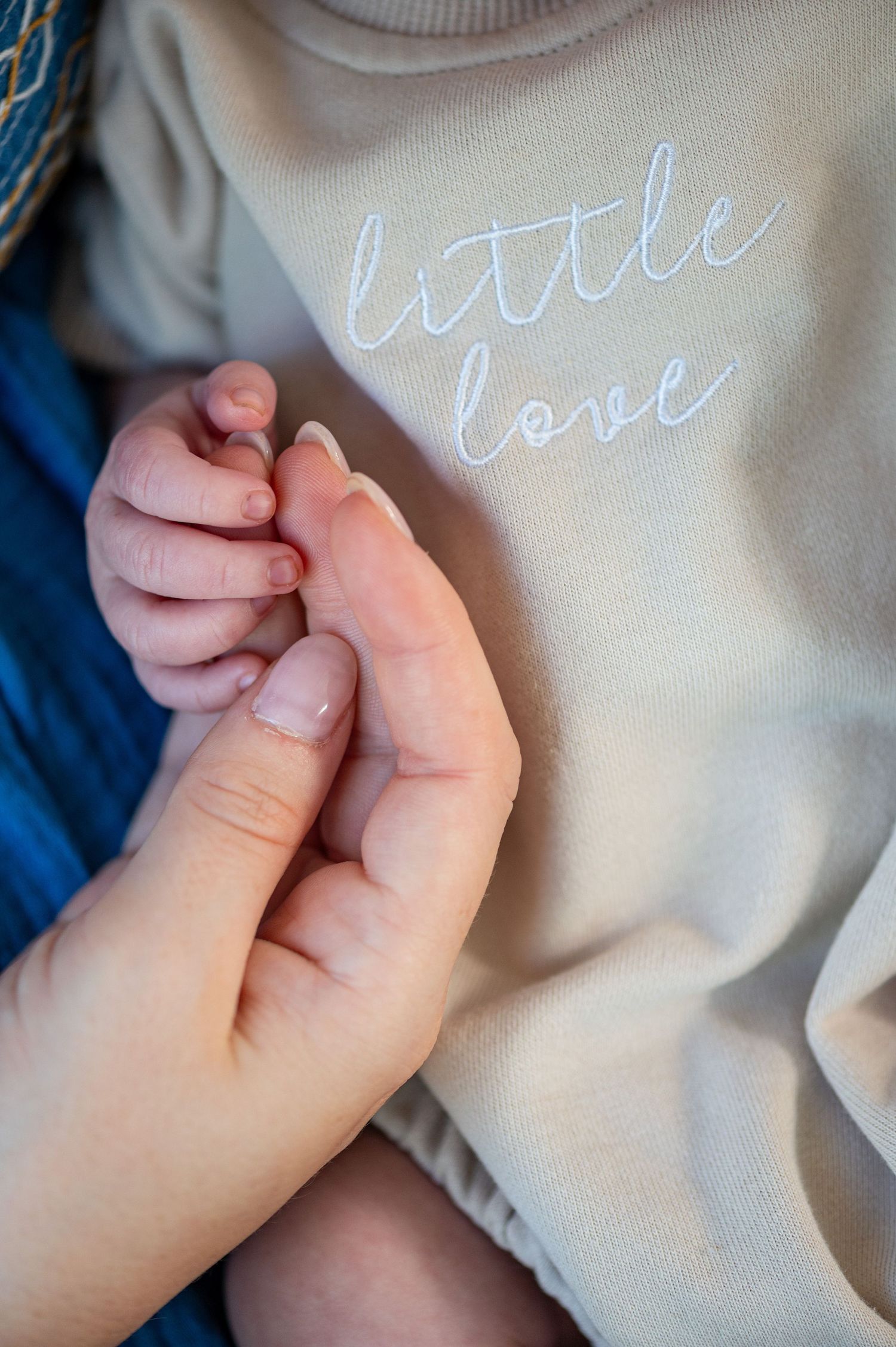 Baby's tiny hand holds onto parent's finger against a light gray sweater with 'little love' text embroidered on it.