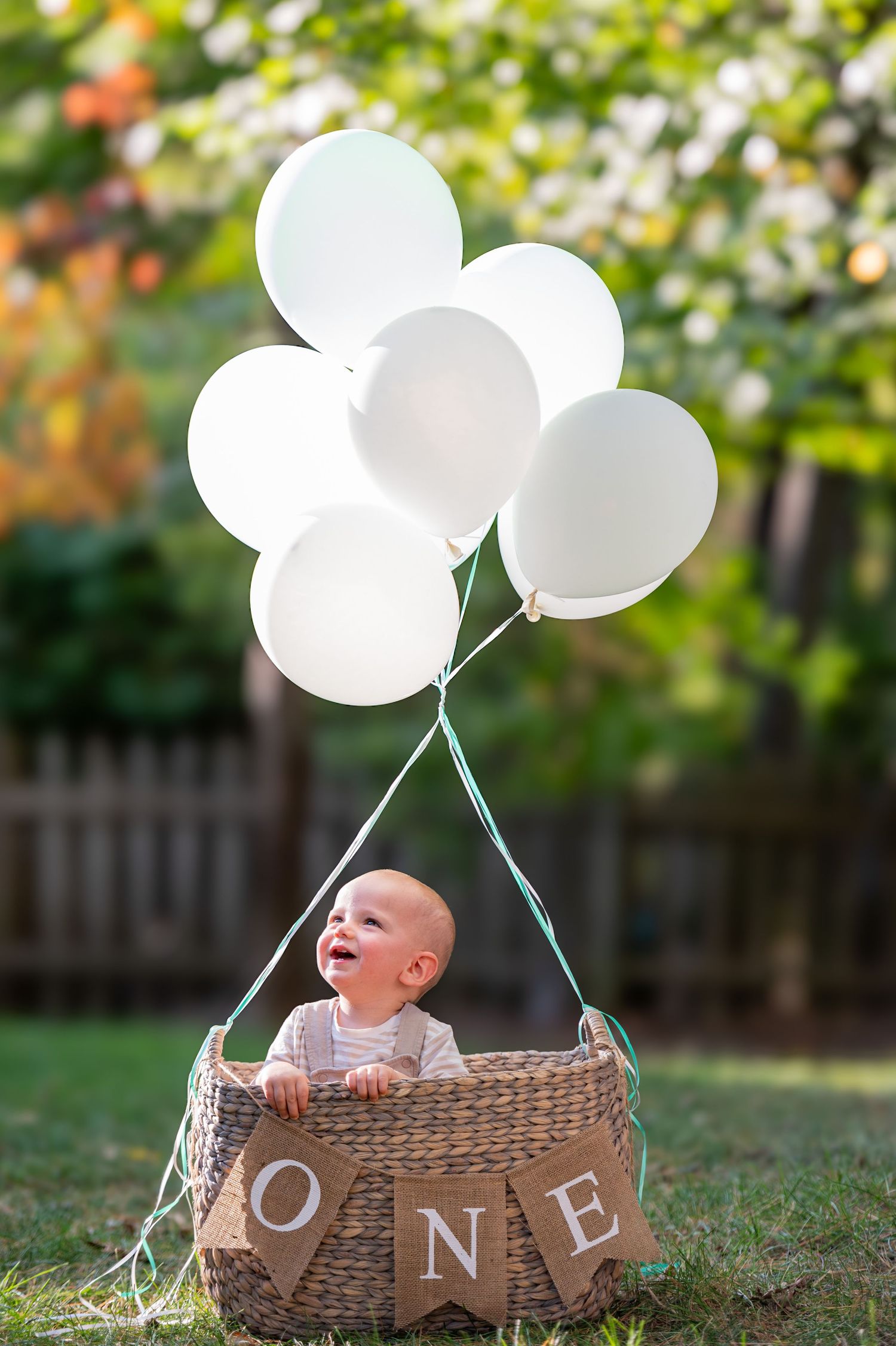 White balloons lift a woven basket with ONE lettering in an outdoor garden setting during a festive celebration.