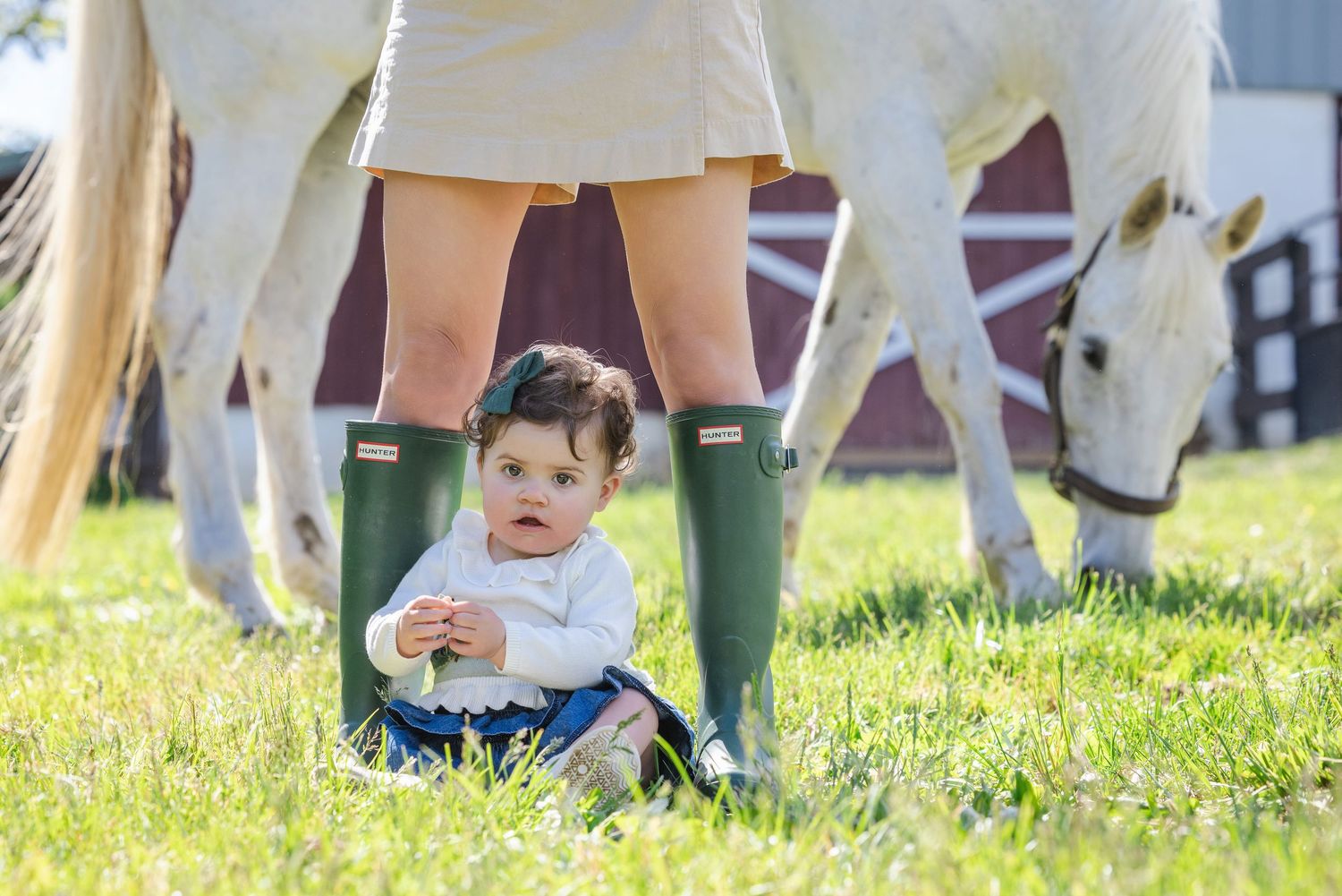 Baby girl sitting between mother's green Hunter boots on grass with white horses grazing near a red barn.
