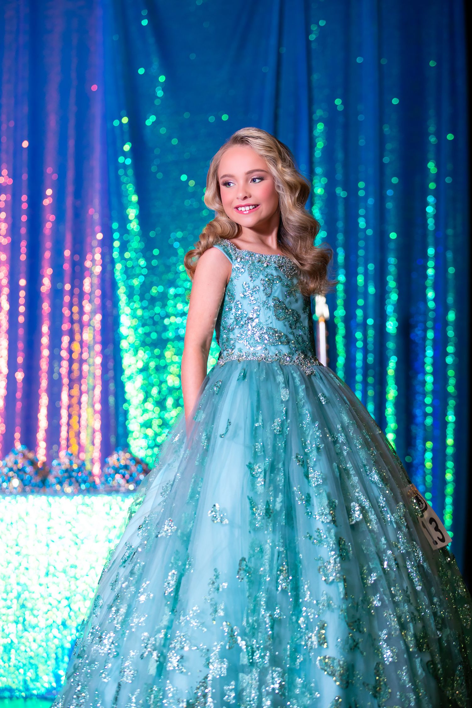 Young girl in a sparkly aqua ball gown on stage with glittering backdrop.