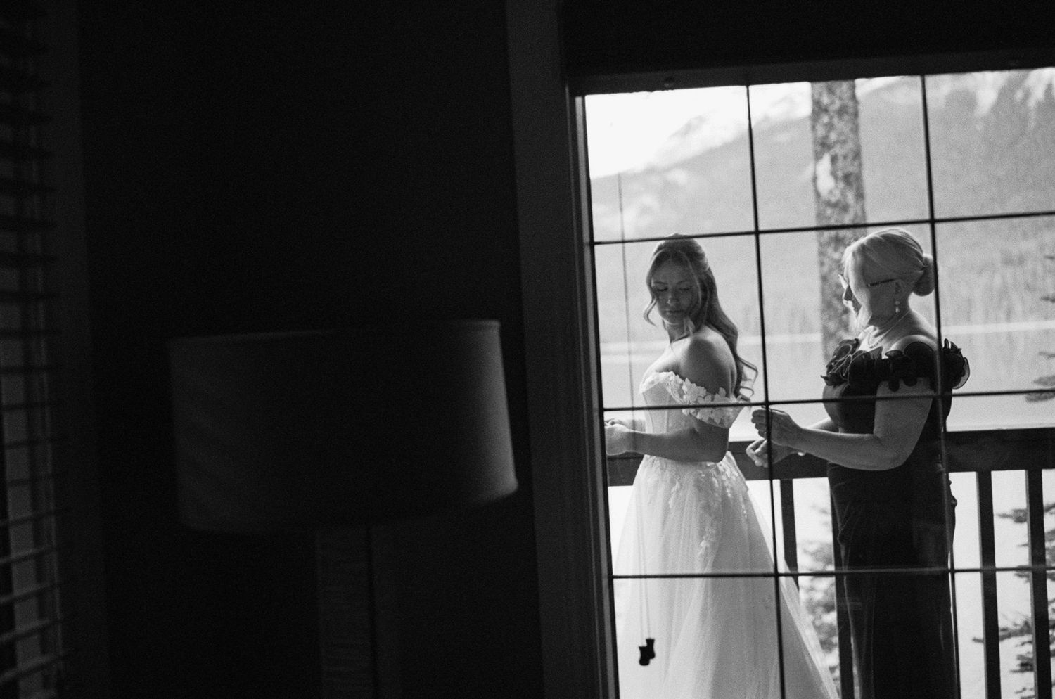 Black and white silhouette of a couple getting ready by a window with mountain views in the background.